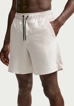 Personne à la peau foncée portant un short blanc avec cordon de serrage et poches latérales, une main dans la poche, debout devant un fond uni.