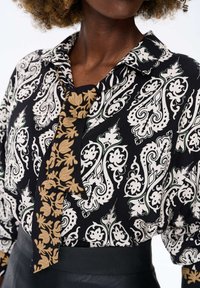 Vrouw in een zwart-wit gestreepte blouse met een contrasterende zwart-tan bloemenstrik en een zwarte hooggerimde rok.