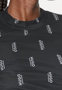 Gros plan d'une personne à la peau foncée portant un t-shirt noir avec des logos blancs "HUGO" répétés dans différentes orientations.