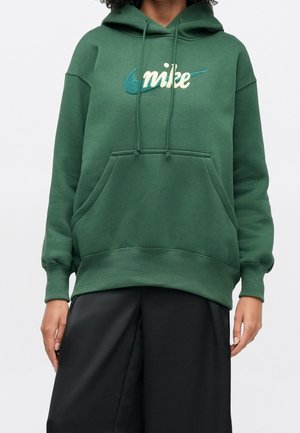 Person iført en grøn Nike hættetrøje med forreste lomme og snore, kombineret med løse sorte bukser, mod en ensfarvet baggrund.
