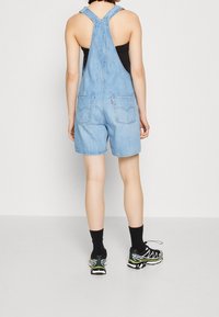 Salopette di denim blu chiaro con bretelle regolabili, gambe corte e due tasche posteriori; abbinata a calzini neri e scarpe da ginnastica.