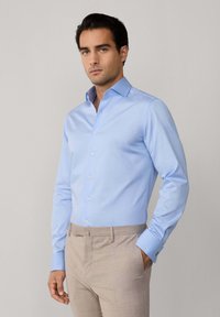 Camicia azzurro chiaro con discreto motivo a reticolo, maniche lunghe e polsini abbottonati. Abbinata a pantaloni beige chiaro, texture liscia e vestibilità su misura.