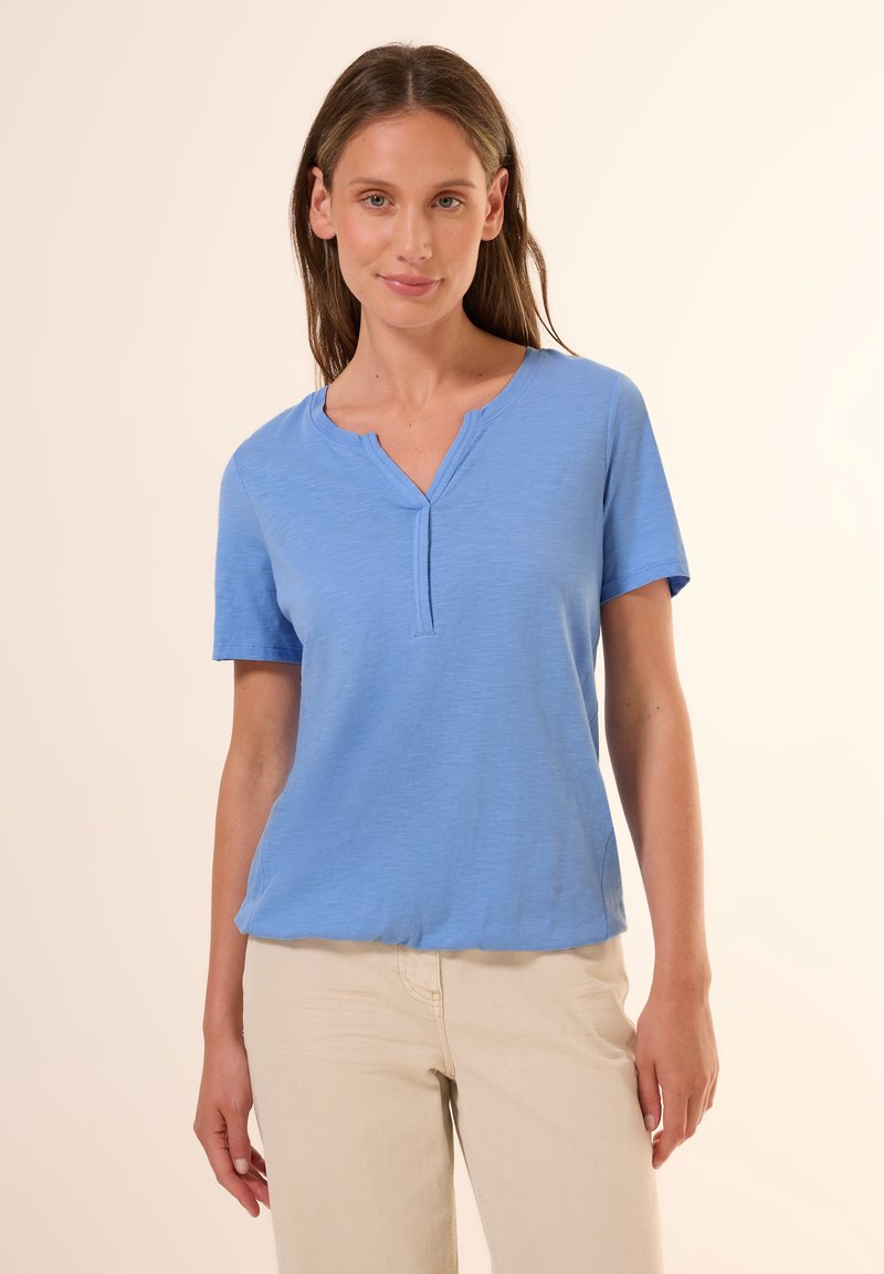 Camicia azzurro chiaro a maniche corte realizzata in morbido tessuto. Design con scollatura a V e un sottile pattina, con una vestibilità rilassata. Abbinata a pantaloni color crema.