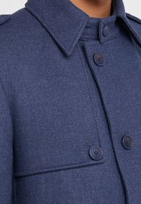 Cappotto di lana blu navy con texture a spina di pesce, colletto strutturato, due grandi bottoni e dettaglio della tasca sul petto.