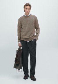 Helles braunes Strickoberteil mit Rundhalsausschnitt, graue Wollhosen und dunkelbraune Schuhe. Eine braune Tasche und ein Schal hängen an einem Arm.