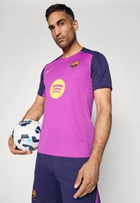 Muž v purpurovom a námorníckom futbalovom drese FC Barcelona, ktorý drží biely a modrý futbalový loptu Nike na neutrálnej pozadí.