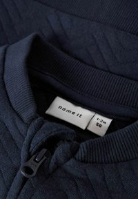 Námořnická zipová bunda vyrobená z texturované pleteniny s límečkem, s malou nálepkou, která zobrazuje značku "name it" a informace o velikosti.