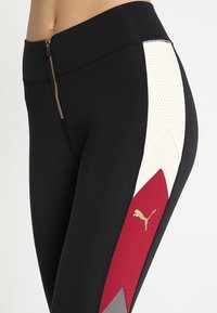 Leggings neri con zip laterale, caratterizzati da un pannello testurizzato bianco e un design a zig zag rosso. Accento con logo dorato sulla parte bassa della gamba.