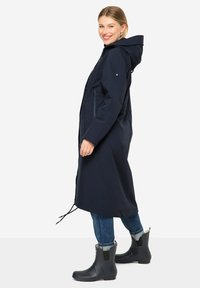 Cappotto lungo della marina con cappuccio, due tasche laterali, vita regolabile con cordino e materiale impermeabile. Indossato sopra jeans blu e stivali da pioggia neri.