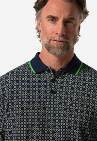 Chemise polo à manches courtes bleu marine avec un motif géométrique multicolore, des accents de col verts et une patte de boutonnage à trois boutons. Détail de tissu texturé visible.