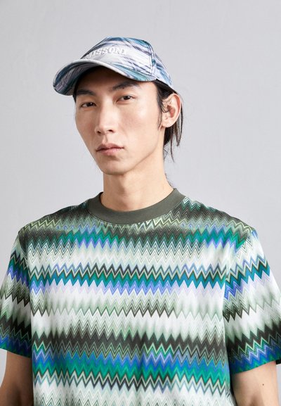 T-shirt à motif zigzag multicolore en vert, bleu et blanc ; associé à une casquette bleu clair avec un logo sur le devant.