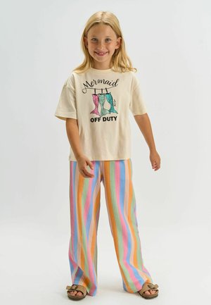 Naeratav tüdruk, kes kannab kreemja värvi t-särki tekstiga „Mermaid Off Duty“ ja kolme värvilist merineitsi saba, triibulisi pastelseid pükse ja sandaale.