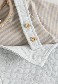 Gestreiftes Oberteil in Beige und Grau mit einem runden Halsausschnitt. Grau gestepptes Latzkleid mit umgeschlagenen Bündchen und Holzknöpfen an den Trägern.