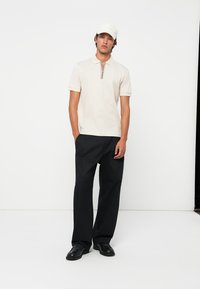 Ung mann som har på seg en beige kortermet polo med glidelås, svarte bredbeinbukser, svarte sko og en hvit caps, stående mot en hvit bakgrunn.