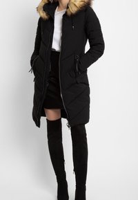 Mujer con abrigo largo negro acolchado y capucha con borde de piel, falda negra, camisa blanca y botas negras por encima de la rodilla, de pie con las manos en los bolsillos.