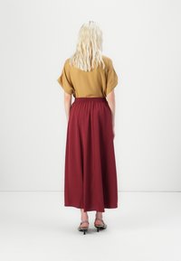 Femme blonde debout, de dos, portant un haut à manches courtes moutarde, une longue jupe bordeaux et des escarpins gris à talons bas sur fond blanc.