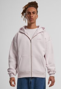 Felpa leggera rosa chiaro con zip, realizzata in un tessuto morbido, dotata di un ampio cappuccio, tasche a marsupio, polsini a costine e un logo ricamato discreto sul petto.