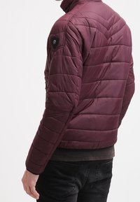 Vínově červená prošívaná bunda s stojatým límcem, horizontálními stehy, kapsou na zip a logem na rameni.
