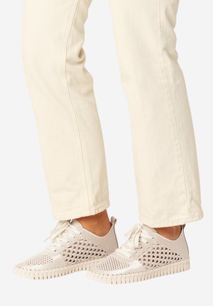 Beige Strick-Sneaker mit Aussparungsmustern, kombiniert mit offwhite-farbenen, gerade geschnittenen Hosen, sichtbar ab der Mitte der Wade abwärts.