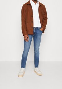 Chaqueta marrón de botones, camiseta polo blanca, vaqueros azules, sneakers crema. La chaqueta tiene cuello y bolsillos frontales; los vaqueros son ajustados.