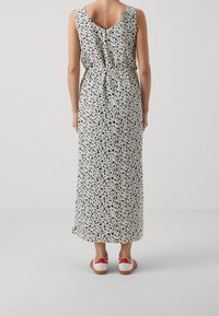 Maxi jurk in crème met een zwart en blauw luipaardprint, voorzien van een V-rug, een taille die is geaccentueerd, en gecombineerd met witte schoenen met rode accenten.