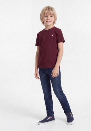 Lyshåret dreng står og smiler iført en burgundy t-shirt, mørkeblå jeans og marineblå slip-on sko mod en hvid baggrund.