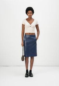 Kurzärmliges, cremefarbenes Oberteil mit Twisted-Detail, kombiniert mit einem dunkelblauen Leder-Bleistiftrock. Das Model trägt schwarze Schuhe und hat eine kleine Tasche dabei.