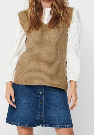 Kobieta ubrana w białą bluzkę z długimi rękawami i falbankami, brązową dzianą kamizelkę bez rękawów oraz niebieską spódnicę dżinsową zapinaną na guziki z przodu.