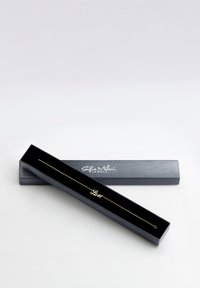 Guldbestick med ordet "Kärlek" i handstil, presenterad i en svart box med slät yta och logotyp på locket.