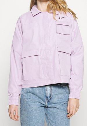 Henkilö, jolla on päällään vaaleanvioletti takki, jossa on useita etutaskuja ja musta Nike-logo, yhdistettynä vaaleansinisiin farkkuihin.