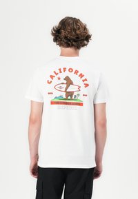 Hvid bomulds t-shirt med et farverigt grafik af en brun bjørn, der holder et surfbræt, med tekstelementerne "CALIFORNIA" og "RIPCURL".
