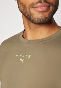 Olive roheline sportlik särk ümmarguse dekoltee ja neonkollase tekstiga "HYROX" ning väikese logoga. Sile ja veniv kangas.