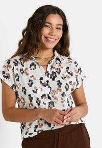 Chemise à manches courtes et à boutons présentant un fond blanc avec un imprimé léopard multicolore dans des tons de marron, noir et accents dorés.