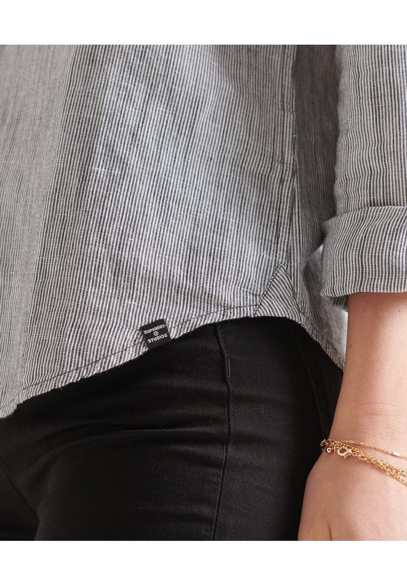 Camicia a righe bianco e nero con maniche arrotolate, tessuto testurizzato e un piccolo etichetta del marchio sull'orlo, abbinata a pantaloni neri.