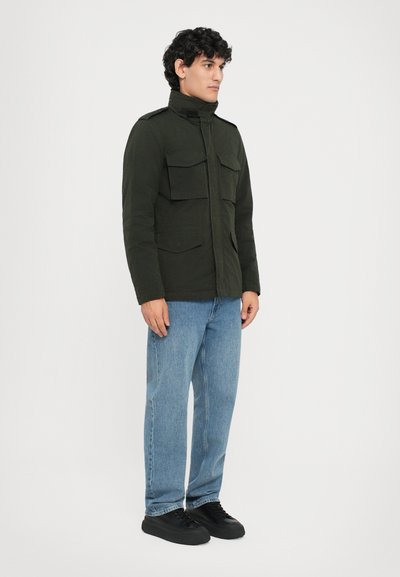 Militärstil-Jacke in Dunkelgrün, mit vier Vordertaschen, hohem Kragen und geradem Schnitt, kombiniert mit hellblauen Jeans und schwarzen Schuhen.