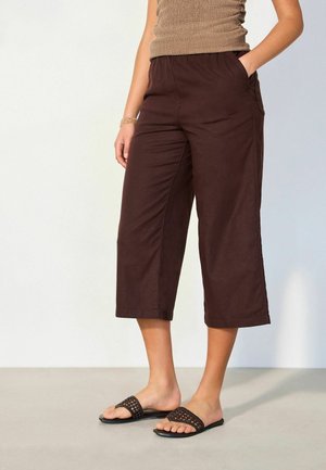 Person wearing dark brown cropped wide-leg pants, tan sleeveless top, and black woven slide sandals, standing with one hand in pocket.