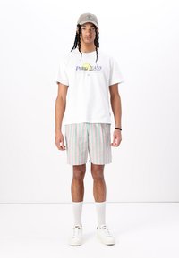 T-shirt en coton blanc avec logo bleu et jaune, associé à un short rayé rose et vert. Porté avec des baskets blanches et une casquette grise. Chaussettes blanches.