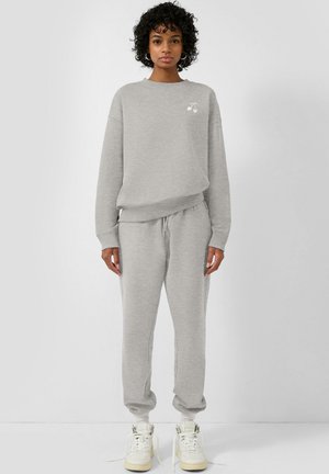 Femme debout portant un sweat-shirt gris clair avec un petit motif de cerise blanche et un pantalon de survêtement assorti, associé à des baskets montantes blanches.