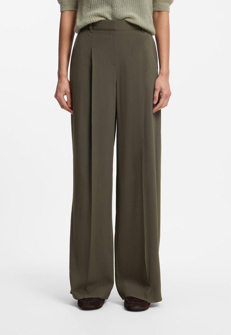 Personne portant un pantalon taille haute à jambes larges vert olive et un pull en maille vert clair, debout les mains relâchées le long du corps.