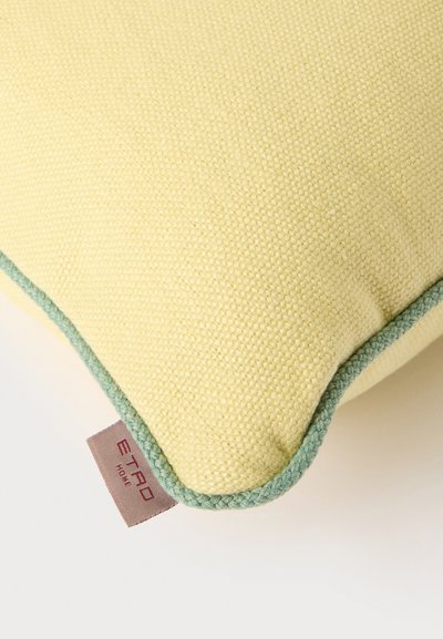 Coin d'un coussin en tissu jaune texturé avec une bordure tressée verte et une petite étiquette marron portant la mention "ETRO HOME."