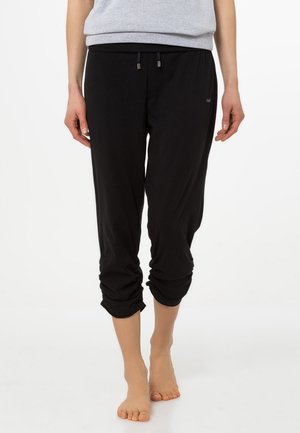 Persoon draagt zwarte cropped joggerbroek met geplooide boorden en een grijze top, staande op blote voeten tegen een witte achtergrond.