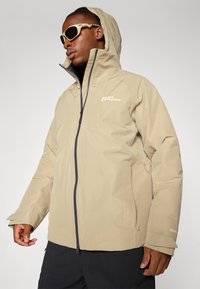 Beige vandtæt jakke med lynlås foran, hætte og justerbare manchetter. Har et logo på brystet og lynlåslommer på siden.
