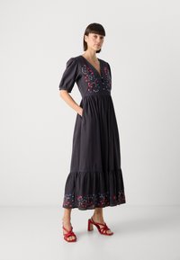 Dunkelgraues Kleid mit bestickten Blumenmustern in Rot und Blau, kurzen Puffärmeln, V-Ausschnitt, gestuftem Saum und Seitentaschen. Kombiniert mit roten Sandalen.