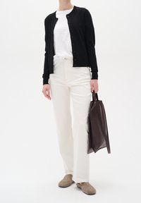 Person som bär en svart knappad kardiagan, vit t-shirt, krämfärgade byxor, beigefärgade loafers och håller en mörkbrun läderhandväska.