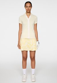 Koszulka polo w jasnym żółtym kolorze z kołnierzykiem i zamkiem błyskawicznym, zestawiona z plisowaną żółtą spódnicą z subtelnym abstrakcyjnym wzorem.
