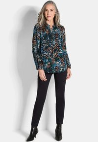 Blusa a maniche lunghe con motivo in teal, nero e marrone, abbinata a pantaloni neri slim-fit e stivali neri con tacco. Tessuto testurizzato.