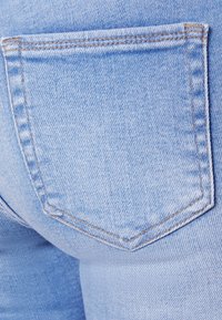 Jeans de denim azul claro com um bolso traseiro retangular com costura que contorna a sua forma, destacando uma superfície suave e texturizada.