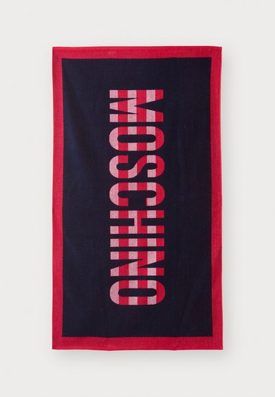 Serviette de plage rectangulaire bleu marine avec une bordure rouge, arborant un texte rose en gras, vertical, indiquant "MOSCHINO". Texture douce, en matériau coton.