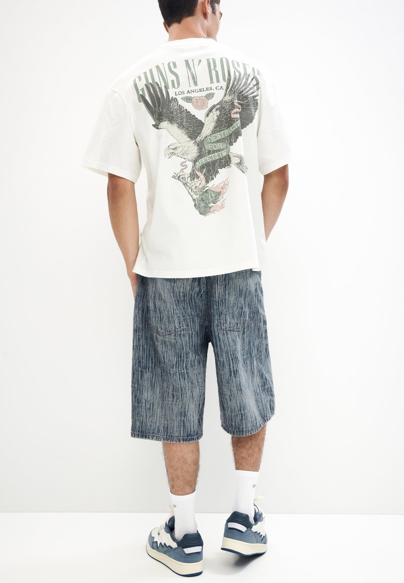 Hvid grafisk T-shirt med ørnedesign og tekst på ryggen; parret med blå strukturerede shorts og hvide sokker, sneakers med blå detaljer.