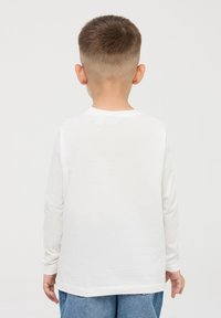 Maglietta bianca a maniche lunghe, realizzata in tessuto morbido, vista da dietro. Presenta un collo rotondo e una vestibilità rilassata, abbinata a jeans blu.